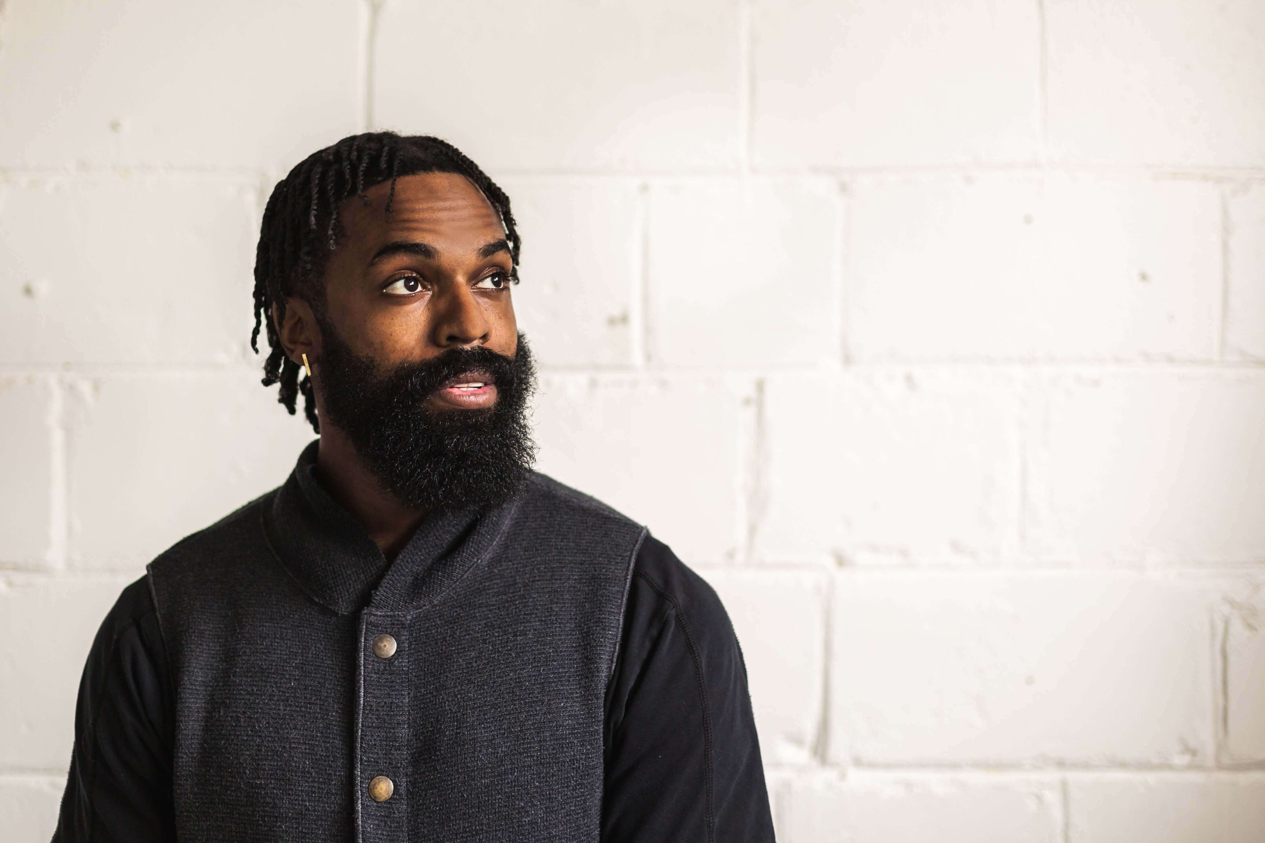 Man with a beard and braided hair wearing a dark jacket against a white brick wall.