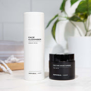 White bottle of face cleanser and black jar of peptide moisturizer on a light surface with a blurred plant in the background.
