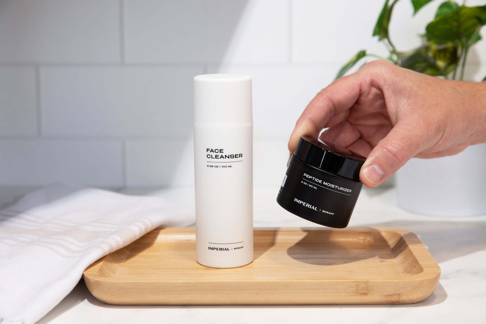 Two skincare products on a wooden tray with a hand interacting with one of them, against a bathroom background.